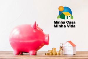 Imagem de cofrinho ao lado de casa em miniatura, ilustrativa para texto sobre o minha casa minha vida.